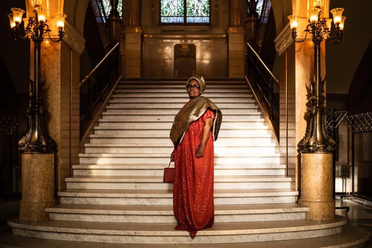 A woman from Krachtvrouwen stands confidently on a grand staircase, wearing a red dress and traditional headwrap in an elegant historic interior.