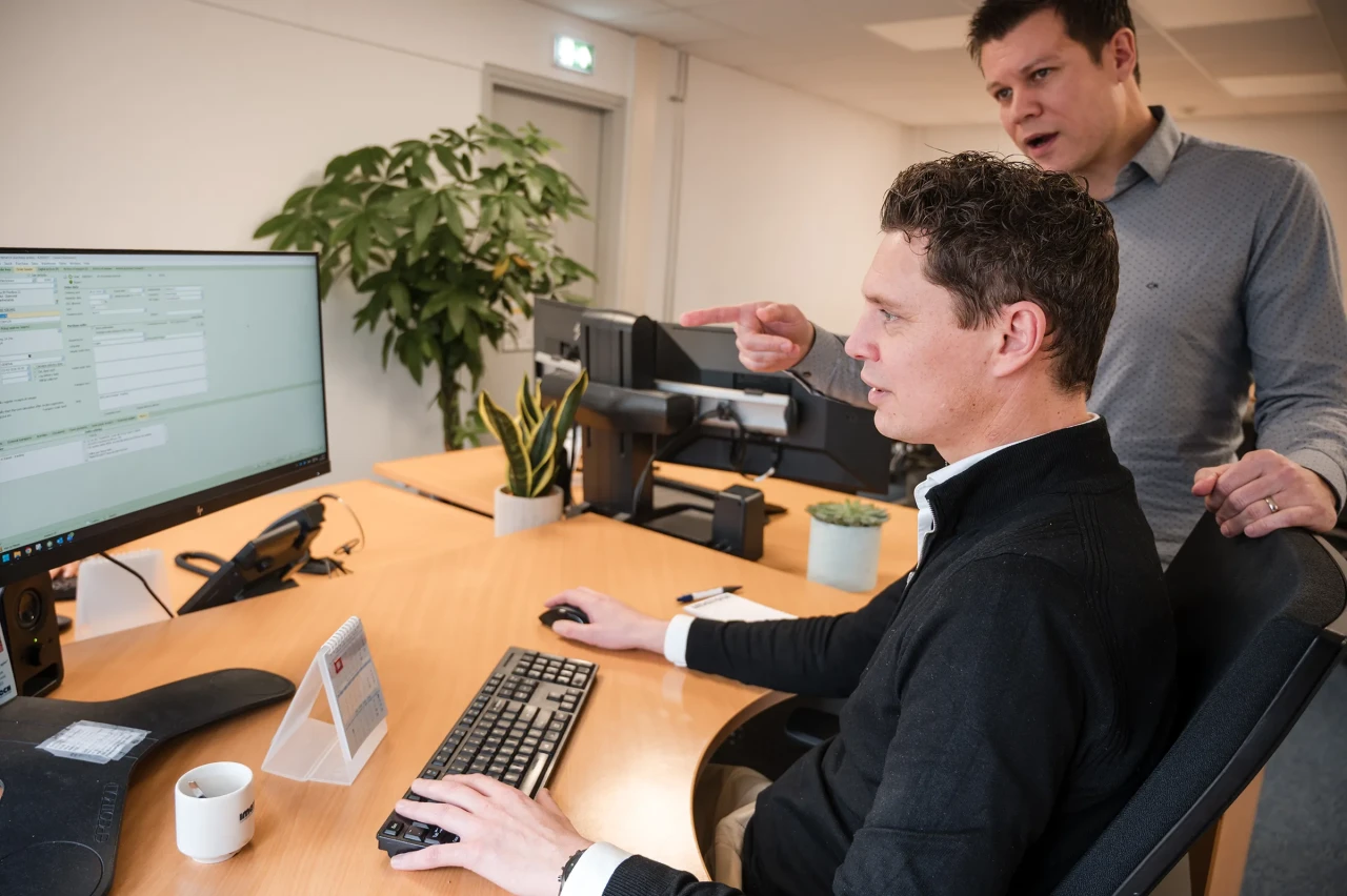 Two Imatech employees review data on a computer screen, as one points out details while the other works at his desk.