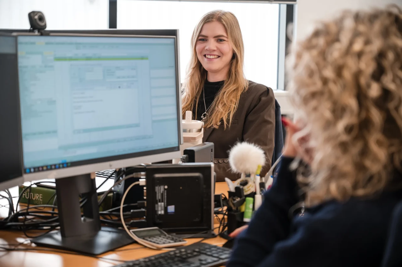 Two colleagues work at adjacent desks, as a woman smiles toward a coworker while computer screens display software in a bright office setting.