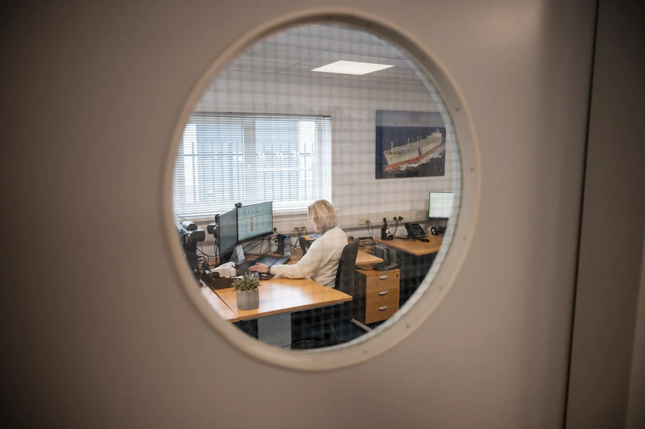 View through a round window shows an Imatech employee working at her desk, handling requests and coordinating order processing in the office.
