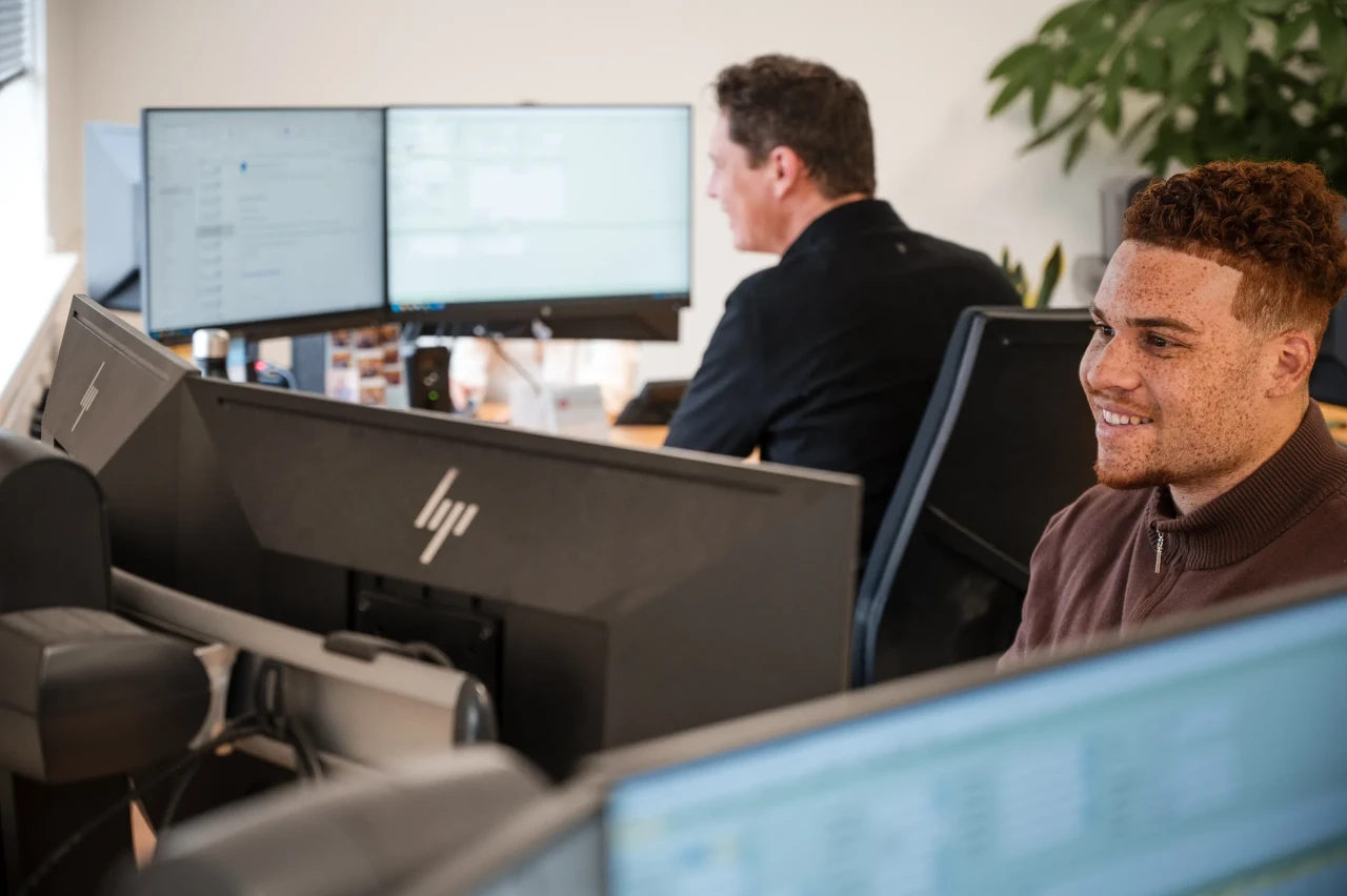 Two colleagues work at computer monitors in a modern office, as one smiles while focusing on his screen.