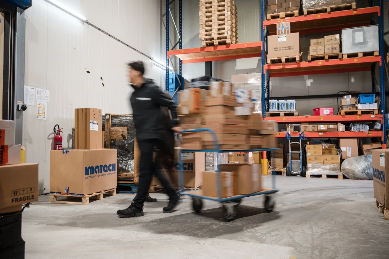 Imatech warehouse employee transporting boxes on a cart through a logistics warehouse with pallets and stored goods.