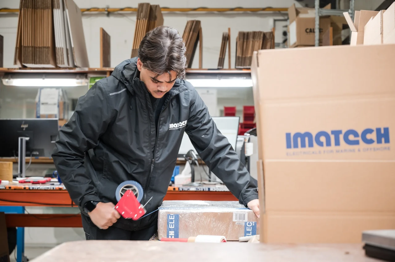An Imatech employee secures a packaged shipment with tape in a warehouse, preparing equipment for delivery beside stacked Imatech boxes.
